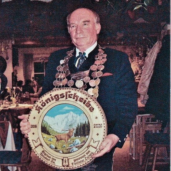 Ein Mann in einem Anzug, der eine Medaille trägt, hält eine runde Platte mit einem Bild eines Hirsches und Bergen vor einem unscharfen Hintergrund.