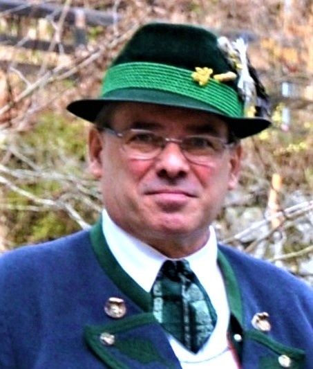 Ein Mann in einer blau-grünen bayerischen Jacke, einem grünen Hut mit einer gelben Blume und einer Brille.