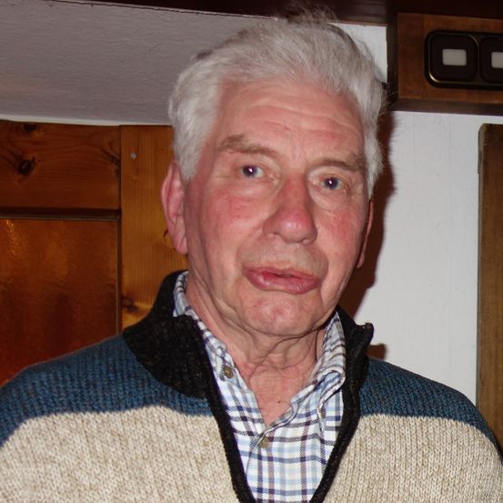 Ein älterer Mann mit weißem Haar und einem Pullover steht vor einer Holztür.