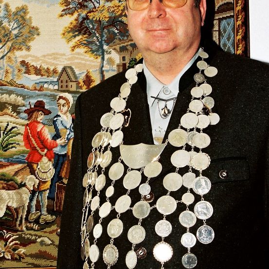 Ein Mann mit Brille und einem schwarzen Anzug steht vor einem Wandteppich. Er trägt eine Kette aus Münzen. Hinter ihm ist ein Gemälde von zwei Menschen und einem Hund.