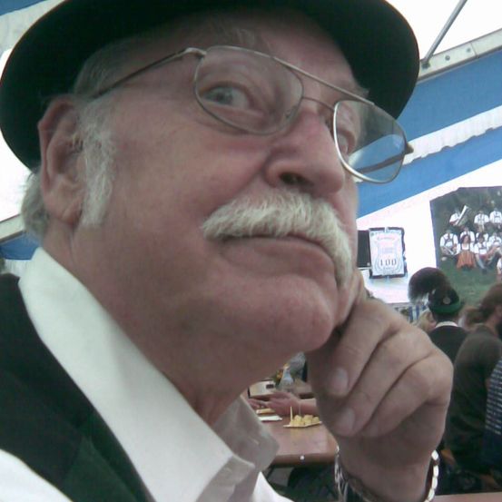 Ein älterer Mann mit Schnurrbart und Brille, der einen schwarzen Hut trägt, sieht nachdenklich aus. Er hält sich mit der rechten Hand das Kinn. Der Hintergrund zeigt ein verschwommenes Bild einer Gruppe von Menschen.