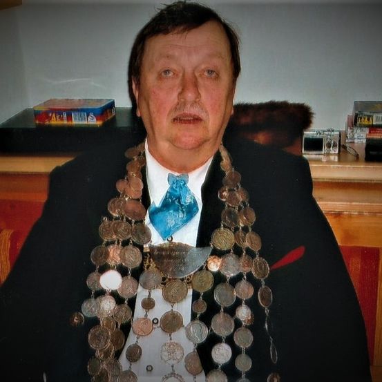 Ein Mann in Anzug und Münzhalskette posiert für ein Foto in einem Raum mit weißer Wand.