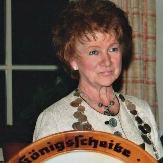 Eine ältere Frau trägt einen weißen Pullover und eine Halskette mit Münzen und steht vor einem hölzernen Schild.