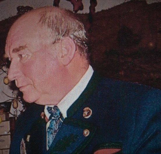 Ein älterer Mann in einem dunkelblauen Anzug und einer Krawatte mit einer Anstecknadel am Revers, der seitlich in einem Raum mit Holzwänden und Schubladen mit hängenden Äpfeln darüber schaut.