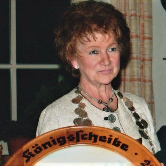 Eine ältere Frau mit kurzen Haaren und einer Halskette lächelt für ein Foto, während sie eine hölzerne Plakette mit der Aufschrift 'Königsstube' hält. Hinter ihr befindet sich ein Fenster mit Vorhängen und ein Topf mit einer Pflanze.