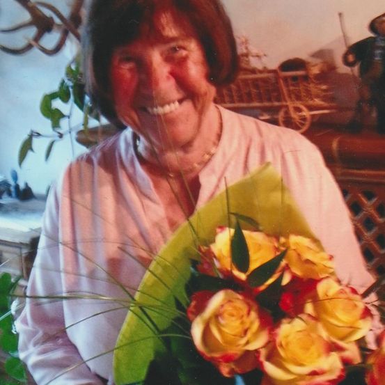 Eine lächelnde Frau hält einen Strauß gelber Rosen und steht in einem Raum mit verschwommenem Hintergrund, der eine Pflanze, einen Holzwagen und dekorative Gegenstände zeigt.