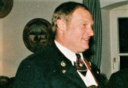 Ein Mann in einem schwarzen Anzug und einer Krawatte mit einer roten Schleife um den Hals lächelt und scheint zu einem Publikum zu sprechen. Hinter ihm befindet sich eine Wand mit einem runden Bilderrahmen und einem Fenster mit grünen Vorhängen.