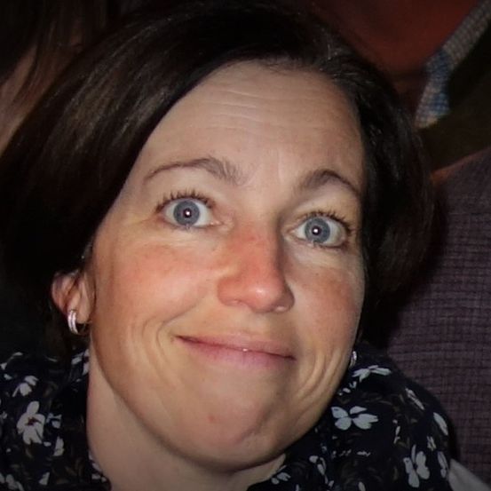 Eine Frau mit braunen Haaren und blauen Augen lächelt mit leicht geöffnetem Mund. Sie trägt ein schwarzes Blumenoberteil. Der Hintergrund ist unscharf.