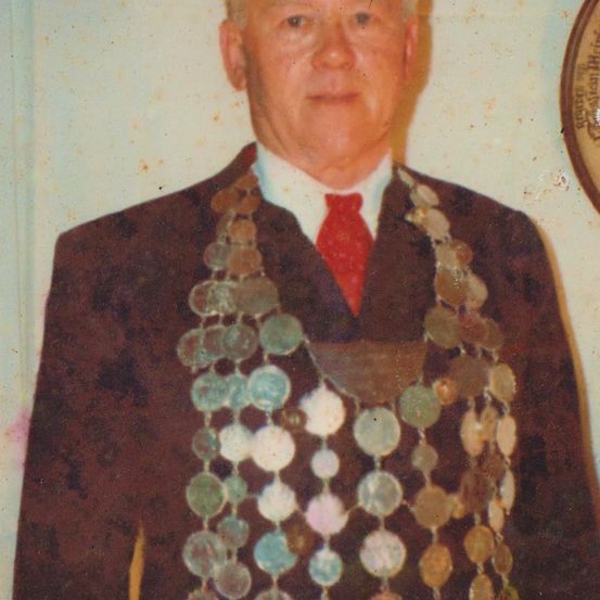 Ein älterer Mann in einem braunen Anzug mit roter Krawatte steht vor einer weißen Wand und trägt eine Halskette aus verschiedenen Münzen.