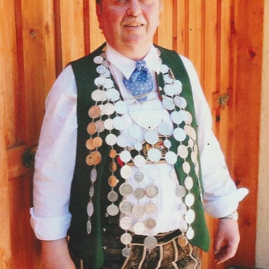 Ein Mann steht vor einer Holztür, trägt eine grüne Weste mit vielen Münzen, ein weißes Hemd, eine blaue Krawatte und traditionelle Hosen.