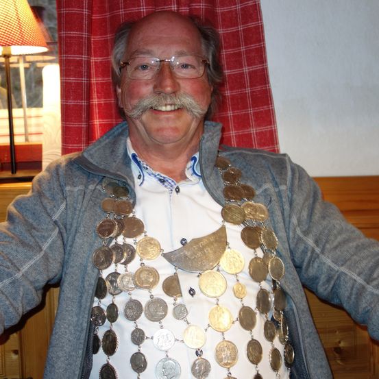 Ein Mann mit Schnurrbart und Brille, der eine Jacke und eine Münzkette trägt, lächelt in einem Raum mit einem roten Vorhang und einer Lampe.