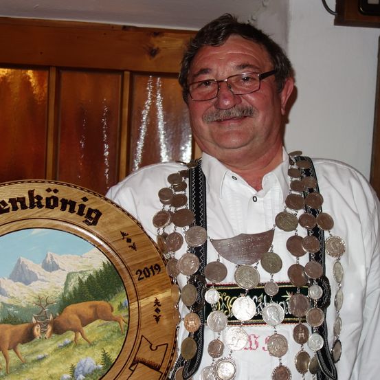Ein Mann mit Brille und Schnurrbart hält eine runde Plakette mit einer Hirschmalerei und '2019'. Er trägt ein traditionelles weißes Hemd mit einer Münzkette.