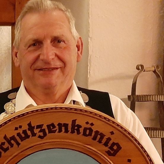 Ein Mann lächelt und hält ein rundes Holzschild mit schwarzem Text. Er trägt ein weißes Hemd, eine schwarze Weste und eine Halskette.