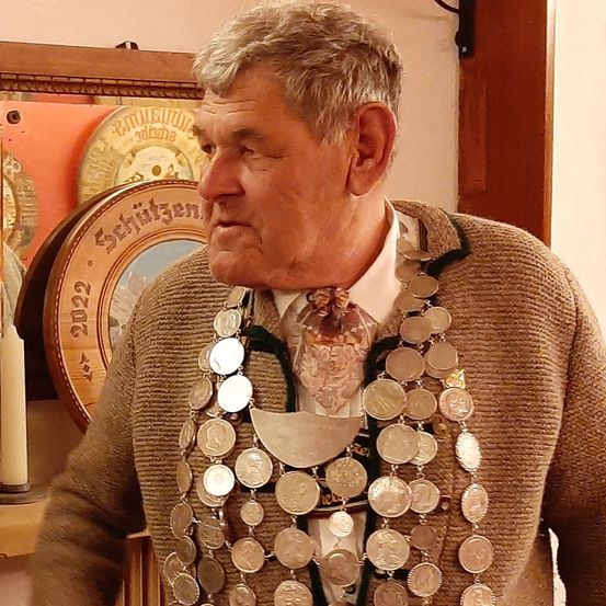 Ein älterer Mann mit grauem Bart und ernster Miene, der einen braunen Pullover und eine Halskette aus Silbermünzen trägt, steht vor einer Wand.