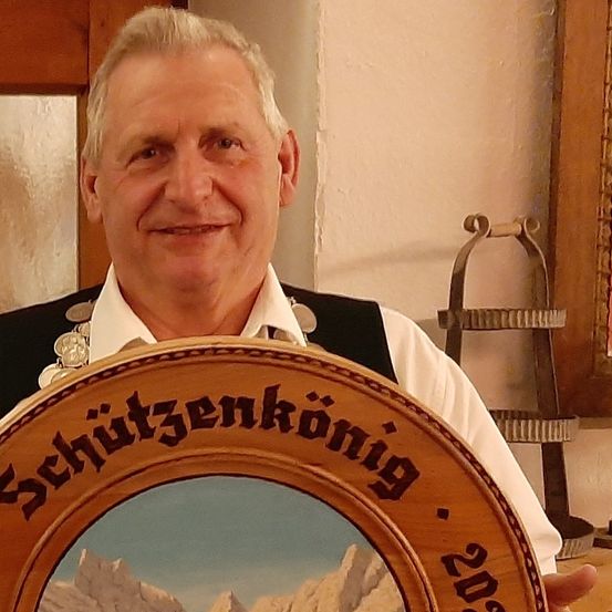 Ein Mann hält eine hölzerne Plakette mit den Worten Schutzenkonnig 2016 vor seiner Brust. Er trägt ein schwarzes Weste und weißes Hemd. Hinter ihm ist eine Wand mit einer Holztür.
