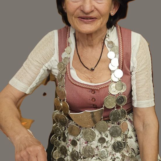 Eine ältere Frau ist in traditioneller Kleidung gekleidet, mit einem rosa Oberteil, das mit einer Silbermünzenschnalle verziert ist. Sie sitzt, lächelt und trägt eine Halskette mit Silbermünzen.