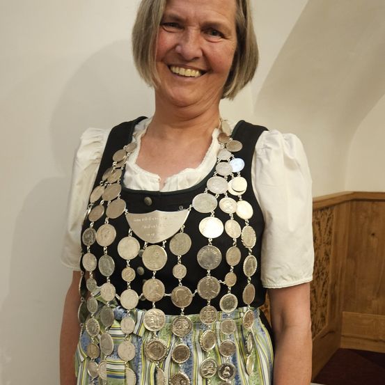 Eine Frau in traditioneller Kleidung mit einer Kette aus Münzen steht vor einer weißen Wand. Sie lächelt.