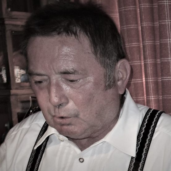 Ein älterer Mann mit einem weißen Hemd und schwarzen Hosenträger mit ernster Miene schaut nach unten. Er hat graues Haar und einen Schnurrbart. Hinter ihm ist ein Vorhang mit kariertem Muster.