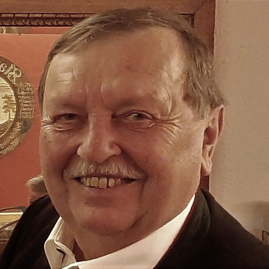 Ein Nahaufnahme eines älteren Mannes, der lächelt, mit einem Schnurrbart und kurzen Haaren, der einen schwarzen Anzug und ein weißes Hemd trägt. Hinter ihm ist eine Wand mit einem dekorativen runden Emblem.