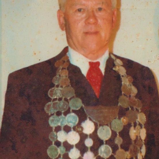 Ein älterer Mann steht mit ernster Miene, trägt einen dunklen Anzug, ein weißes Hemd und eine rote Krawatte. Er trägt auch eine dekorative Kette aus Münzen um den Hals.