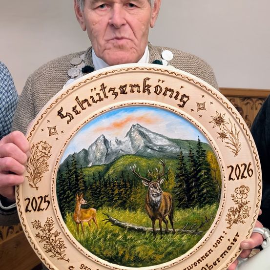 Ein älterer Mann hält einen runden Teller mit einer malerischen Szene von Rehen in einer Berglandschaft, auf der die Jahre 2025 und 2026 eingraviert sind.