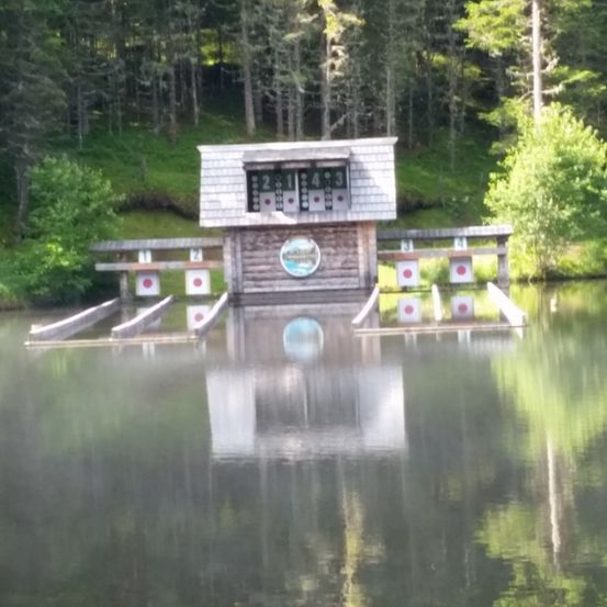 Ein kleines Holzgebäude mit einer Anzeigetafel befindet sich in der Mitte eines Sees, umgeben von Bäumen und Grün. Das Gebäude hat einen runden Spiegel an der Vorderseite und rote und weiße Ziele auf beiden Seiten.