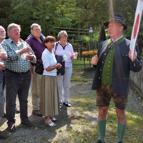 Eine Gruppe von Menschen steht im Freien, ein Mann in traditioneller Kleidung spricht zu ihnen. Einer hält ein Ziel, andere beobachten. Bäume und Grünpflanzen sind im Hintergrund.