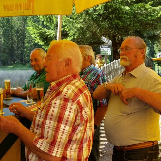 Eine Gruppe älterer Männer sitzt unter einem gelben Schirm an einer Bar und genießt Bier. Der Mann im karierten Hemd unterhält sich mit einem anderen Mann, der eine Brille trägt.
