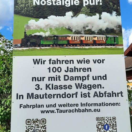 Ein Schild zeigt eine Dampflokomotive auf den Gleisen, die weißen Rauch ausstößt. Es steht 'Nostalgie pur!' und 'Wir fahren wie vor 100 Jahren nur mit Dampf und 3. Klasse Wagen.' Der Abfahrtsort ist Maunterndorf. Weitere Informationen finden Sie unter www.Taurachbahn.eu. Zwei QR-Codes sind vorhanden.