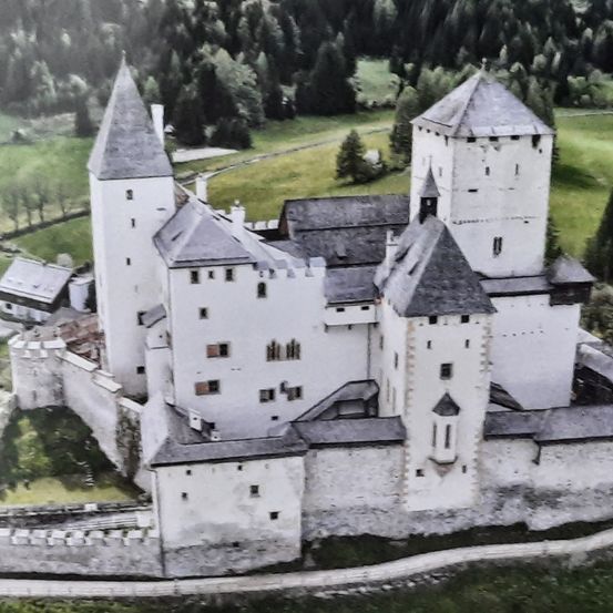 Ein Luftbild eines historischen Schlosses mit zwei hohen Türmen und einem großen Innenhof. Das Schloss ist von einer Steinmauer und üppigen grünen Feldern umgeben.