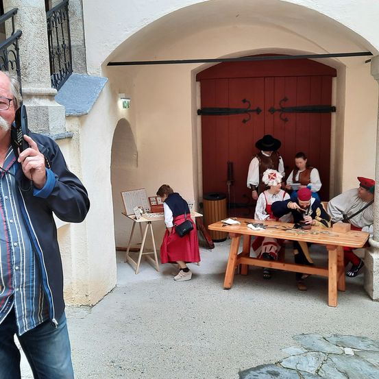 Ein Mann macht ein Foto mit seinem Handy in der Nähe eines historischen Gebäudes, wo mehrere Menschen in historischen Kostümen um einen Holztisch versammelt sind.