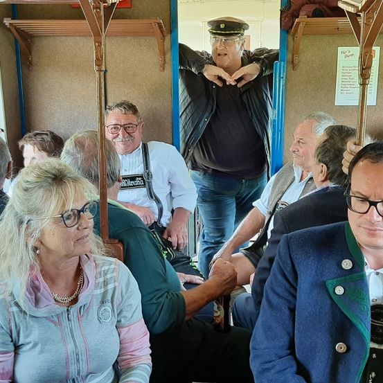 Eine Gruppe von Menschen in einem Bus, ein älterer Mann in einer schwarzen Jacke und einer Mütze steht und macht eine Herzgeste mit den Händen. Eine Frau mit Brille sitzt vorne.