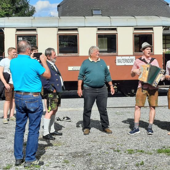 Eine Gruppe von Männern steht vor einem alten Zug, einer spielt Akkordeon. Dahinter trägt der Zug ein Schild mit der Aufschrift 'Mauterndorf.' Die Umgebung ist im Freien, mit Schotterboden und klarem Himmel.