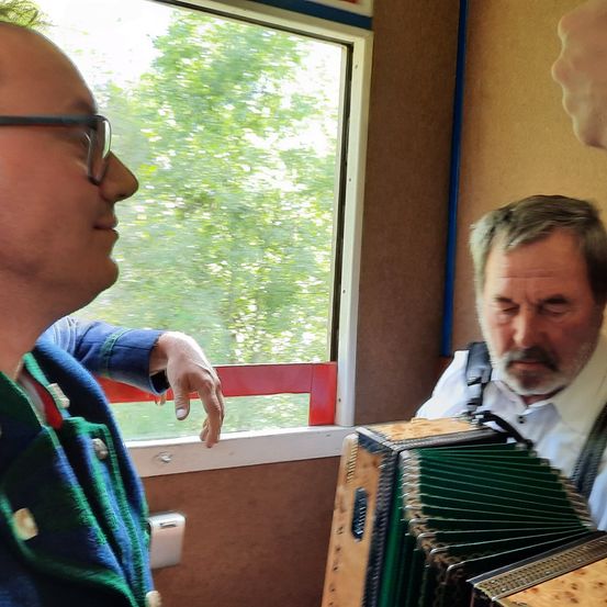 Zwei Männer sind in einem Fahrzeug. Ein Mann spielt Akkordeon, und der andere Mann schaut ihn an. Der Mann, der das Akkordeon spielt, trägt ein weißes Hemd. Das Fahrzeug hat ein Fenster mit Blick auf Bäume.