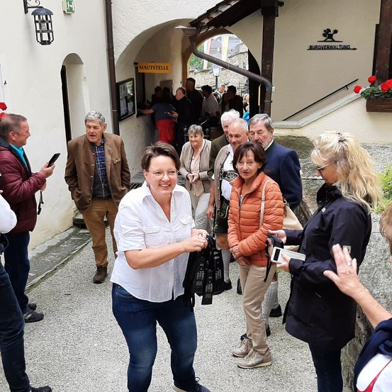 Eine Gruppe von Menschen versammelt sich vor einem Gebäude mit einem Bogeneingang. Die Frau im weißen Shirt lächelt und gestikuliert, während andere Kameras und Taschen halten. Ein Schild über dem Eingang steht 'MAUISTELLE'.