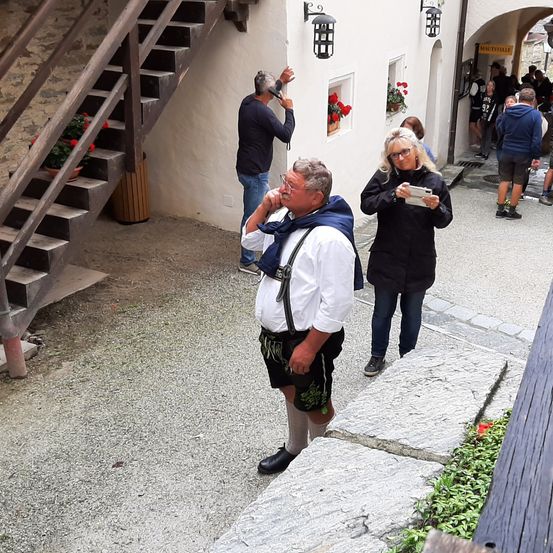 Ein Mann in traditioneller Kleidung steht auf einer Kopfsteinpflasterstraße, während andere um ein weißes Gebäude mit Blumen und Treppen herumlaufen.