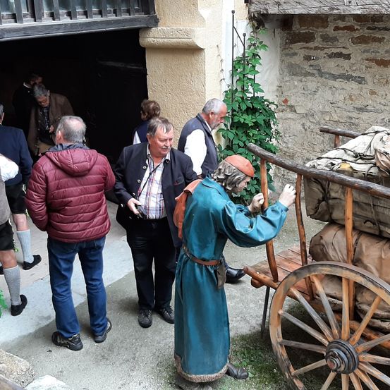 Mehrere Männer in verschiedenen Kleidungsstücken stehen in der Nähe einer Steinmauer, einige schauen auf eine Puppe in historischer Kleidung, die einen Holzwagen mit Säcken hält.