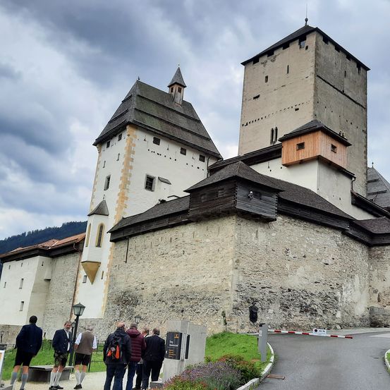 Mehrere Menschen besuchen eine historische Burg mit hohen Türmen und einem bewölkten Himmel im Hintergrund. Sie stehen vor der Burg.