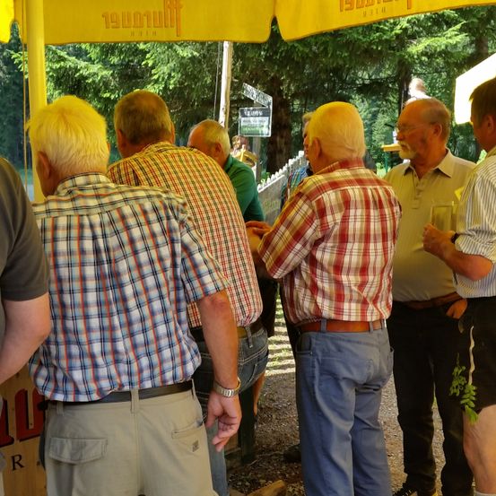 Mehrere ältere Männer versammeln sich unter einem gelben Baldachin. Sie sind in Freizeithemden und Hosen gekleidet. Ein Mann hält ein Glas Wein. Im Hintergrund sind Bäume und ein grünes Schild zu sehen.