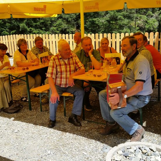 Mehrere Menschen sitzen um Tische unter einem Schirm. Ein Mann in einem karierten Hemd lächelt in die Kamera. Ein anderer Mann spielt Akkordeon. Ein Beutel und ein Haufen Steine sind in der Nähe.
