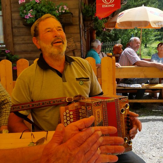 Ein älterer Mann spielt draußen in einem Café ein Akkordeon, umgeben von anderen Gästen.