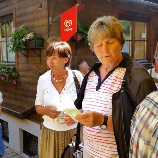 Zwei Frauen stehen vor einem Holzgebäude. Eine liest ein Blatt Papier. Hinter ihnen ist eine rote Fahne und Blumen in einer Fensterbank.