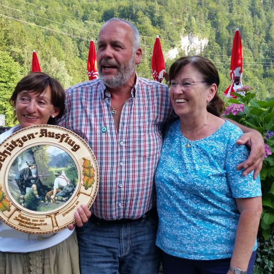 Drei Menschen stehen vor einem grünen Hintergrund und lächeln. Der Mann hat einen Bart und hält einen Teller mit einem Bild darauf. Die beiden Frauen stehen eng beieinander. Dahinter stehen rote Regenschirme.