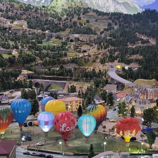 Eine Luftaufnahme einer Modellstadt mit Heißluftballons in verschiedenen Farben und Designs. Gebäude, eine Straße und Berge sind im Hintergrund.