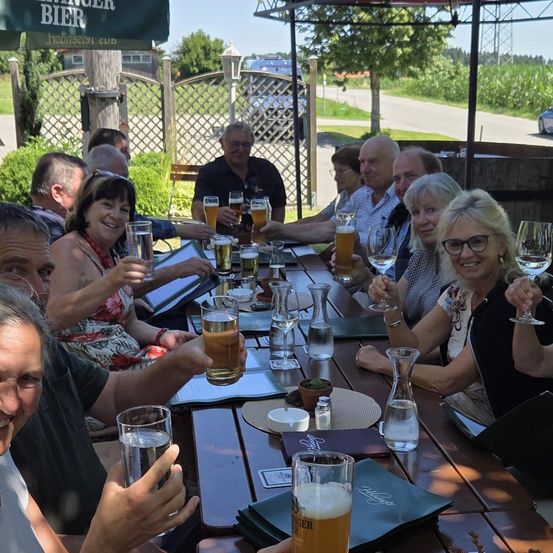 Eine Gruppe von Menschen sitzt um einen Tisch herum und hält Gläser mit Bier in der Hand. Dahinter befindet sich ein Zaun und ein Haus mit einem Schild, das Bierer Bier sagt.