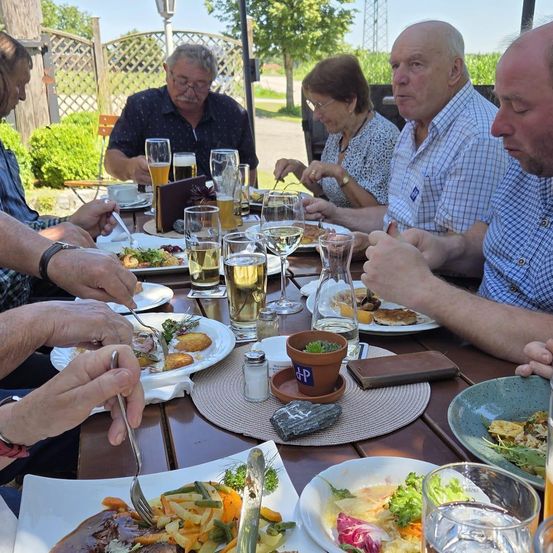 Eine Gruppe von Menschen, die draußen an einem Tisch mit verschiedenen Gerichten, Weingläsern und Bierkrügen essen. Einige tragen Brillen, und es gibt Pflanzen und einen Zaun im Hintergrund.