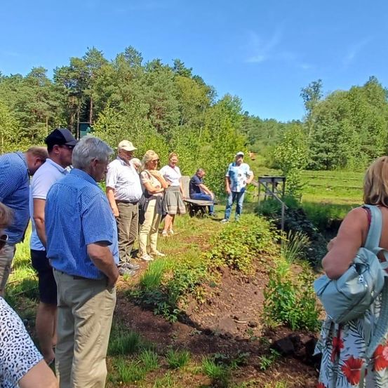 Eine Gruppe von Menschen steht in einem Feld mit Bäumen und Pflanzen. Einige schauen auf etwas am Boden, während andere in Gespräche vertieft sind. Ein Mann mit Hut sitzt auf einer Bank.