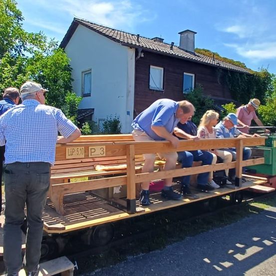 Mehrere Personen sitzen auf einer Holzbank, die an einen Eisenbahnwagen befestigt ist, während ein Mann daneben steht. Dahinter steht ein Haus mit Bäumen und Pflanzen.