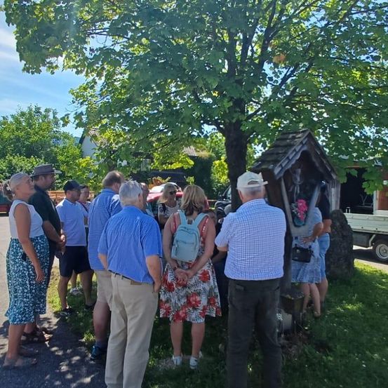 Eine Gruppe von Menschen steht in einem Außenbereich, wahrscheinlich auf einem Bürgersteig. Sie stehen um einen Baum herum, wobei einige von ihnen in Richtung Kamera schauen. Eine Frau steht vor einer kleinen Holzstruktur mit einer Blumendekoration. Ein Lastwagen ist am Straßenrand geparkt.