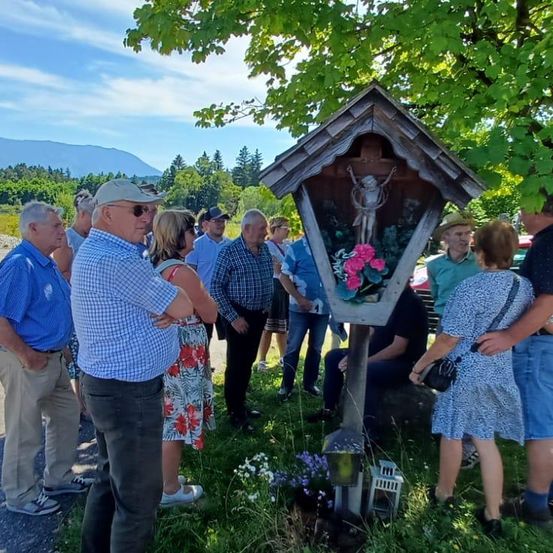 Eine Gruppe von Menschen versammelt sich um ein kleines Wegekreuz mit einem Kruzifix, unter einem Baum, mit Bergen im Hintergrund.